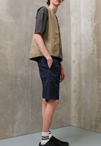 Gilet beige avec poches, t-shirt noir à motif, shorts bleu marine et baskets noires avec rayures blanches sur les chaussettes. Tenue simple et décontractée.