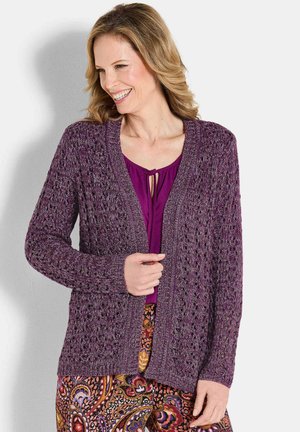 Donna sorridente, che indossa un cardigan viola a maglia aperta sopra una blusa viola e pantaloni dai colori vivaci con motivo paisley, su uno sfondo semplice.