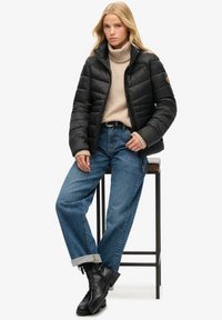 Czarna kurtka puchowa z poziomym przeszyciem, wysoki kołnierz i zamek błyskawiczny, w połączeniu z beżowym golfem i niebieskimi dżinsami. Czarne buty na sznurówki.