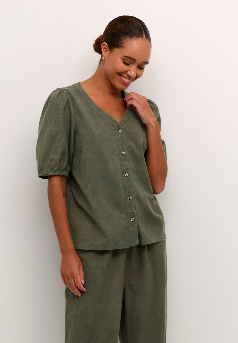 Blouse en lin vert olive avec des manches bouffantes courtes, décolleté en V et boutons sur le devant. Texture lisse, coupe décontractée, s'associe avec un pantalon assorti.