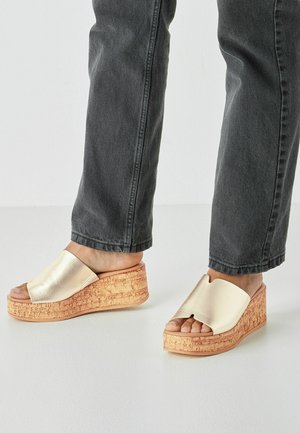 Sandały na koturnie z metaliczną, złotą, syntetyczną wierzchnią częścią i teksturowaną, korkopodobną podstawą. Otwarty nosek bez paska z tyłu.