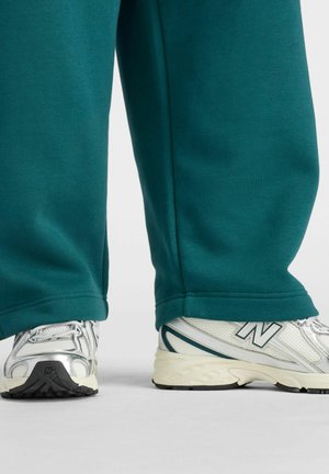 White and silver New Balance sneakers worn with teal wide-legged pants, photographed from the calves down against a plain background.