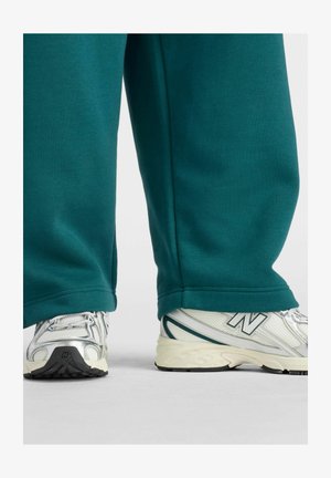 White and silver New Balance sneakers worn with teal wide-legged pants, photographed from the calves down against a plain background.