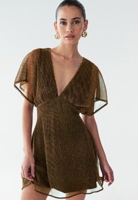 Bronze plisseret kjole med dyb V-hals, gennemsigtige flutterærmer og en flydende silhuet, der har en tekstureret, glitrende overflade.