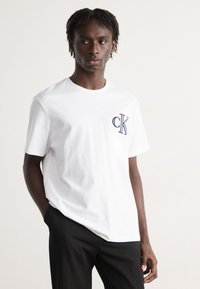 Hvid bomulds t-shirt med et blåt broderet "CK" logo på venstre bryst. Standard rund halsudskæring og korte ærmer.