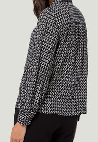 Blouse à manches longues, col et coupe décontractée, à motifs noirs et blancs. Présente un design géométrique répétitif et une texture douce.