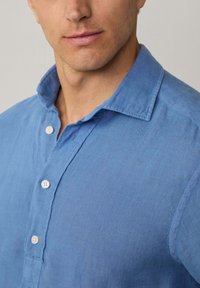 Camicia di lino blu chiaro con colletto con bottoni e patta frontale con bottoni bianchi. Il tessuto ha un aspetto texturizzato e leggermente usurato.