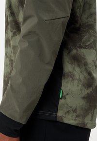 Veste vert olive avec un motif texturé, présentant une manche ajustée, des accents noirs et une petite étiquette verte près de l'ourlet.