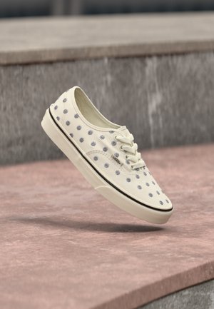 Λευκό αθλητικό παπούτσι Vans με ανοιχτομπλέ λουλουδάτο σχέδιο, λευκά κορδόνια και μαύρη ρίγα στη σόλα, που αιωρείται πάνω από ροζ πέτρινα σκαλοπάτια.