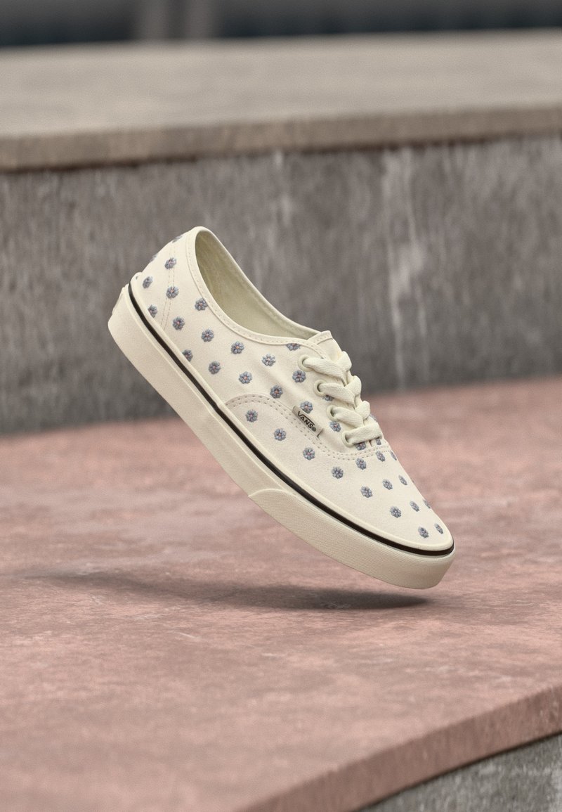 Λευκό αθλητικό παπούτσι Vans με ανοιχτομπλέ λουλουδάτο σχέδιο, λευκά κορδόνια και μαύρη ρίγα στη σόλα, που αιωρείται πάνω από ροζ πέτρινα σκαλοπάτια.