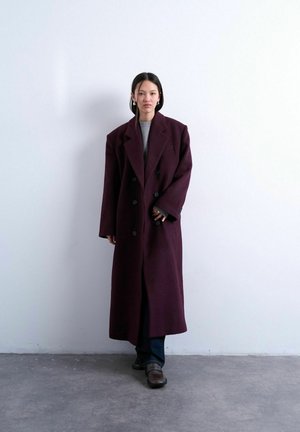 Donna in piedi contro una parete bianca che indossa un cappotto doppiopetto oversize viola scuro, top grigio, pantaloni scuri e scarpe scure.
