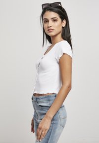 Biely rebrovaný crop top s krátkymi rukávmi, s gombíkovým zapínaním a volánikovými okrajmi, nosený s vysoko pásovými svetlomodrými džínami.