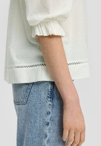 Blouse blanche texturée avec des manches bouffantes et un ourlet froncé, présentant un motif à pois et une finition en dentelle en bas. Portée avec un jean en denim clair.