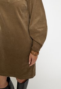 Braunes Cordkleid mit langen Ärmeln, lockerem Schnitt und strukturierter Oberfläche. Ein großer, hellbrauner Armreif schmückt das Handgelenk. Schwarze Stiefel bis zum Knie sind sichtbar.