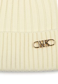 Cremfarbener Strickmütze mit geripptem Muster und goldfarbenem Logo-Detail. Das Design verfügt über einen umgeschlagenen Bund und ein weiches, warmes Material.