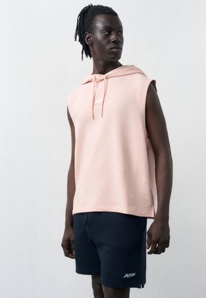 Tumedanahaline mees dreadlockside kannab varjatud roosa varrukateta pusas ja tumesinistes lühikestes pükstes, millel on "BOSS" logo, vaadates kõrvale.