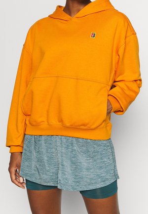 Persona con sudadera con capucha de color naranja brillante con un pequeño logo en el pecho y pantalones cortos deportivos gris claro sobre pantalones cortos de ciclismo verde oscuro.