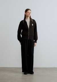 Cardigan marron côtelé avec une fermeture éclair à l'avant, bords de col blancs et petit logo en forme de diamant. Porté avec un pantalon noir ample et des chaussures noires.