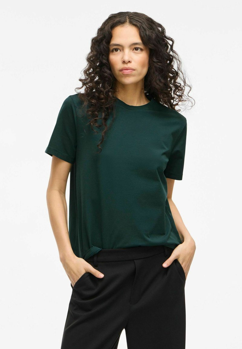 T-shirt verde scuro realizzata in un tessuto morbido, con maniche corte e scollatura tonda classica, indossata con pantaloni neri.