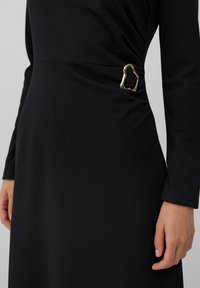 Schwarzes langärmliges Kleid mit einer goldenen, unregelmäßig geformten Schnalle auf der rechten Seite in der Taille.