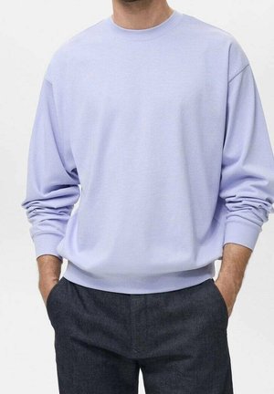 Mann trägt ein hell lavendelfarbenes Sweatshirt mit langem Arm und Rundhalsausschnitt, die Hände in den Taschen einer dunkelgrauen Hose.