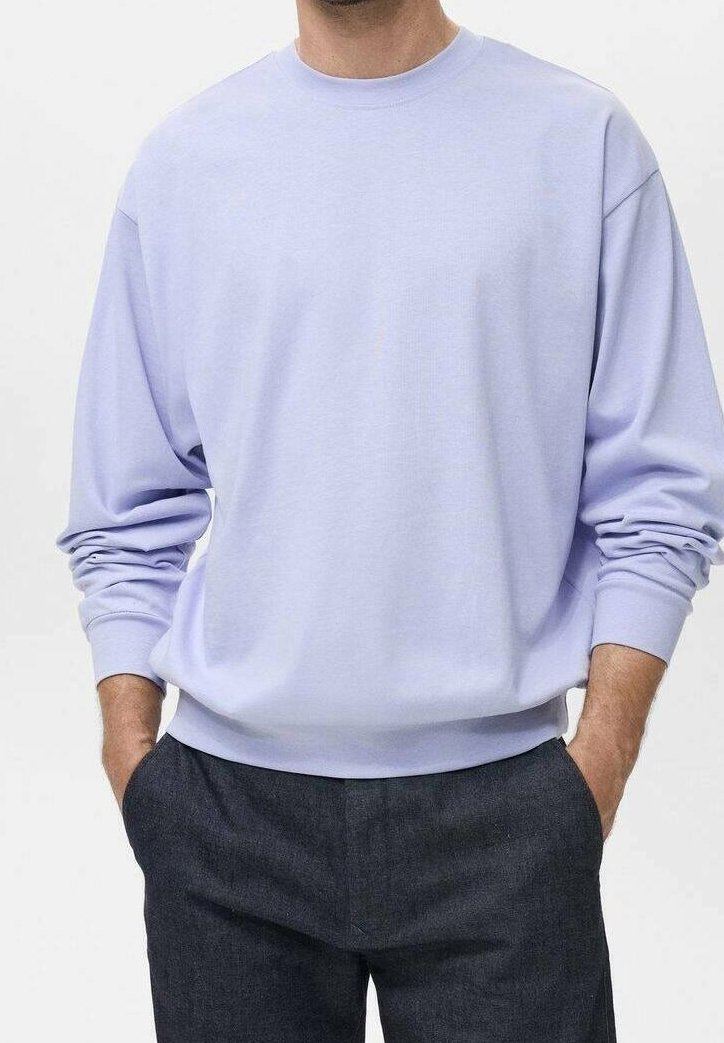Homme portant un sweat-shirt à col rond à manches longues lavande clair, les mains dans les poches d’un pantalon gris foncé.