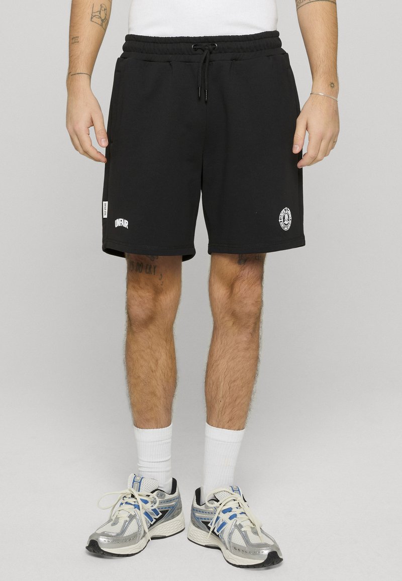 Homme portant un short noir à cordon avec des logos blancs, des chaussettes blanches hautes et des chaussures de course argentées, bleues et noires, debout sur un sol gris clair.