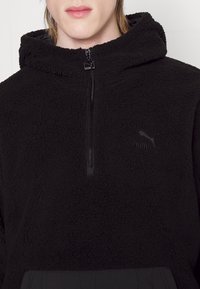 Felpa nera in pile con zip frontale a metà, superficie testurizzata e dettaglio del logo sul petto. Con cappuccio e tasca a marsupio.