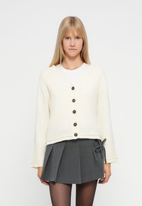 Cremfarbener Cardigan mit V-Ausschnitt, fünf braunen Knöpfen und Glockenärmeln, kombiniert mit einem hoch taillierten grauen Plisseerock. Glatte Textur.