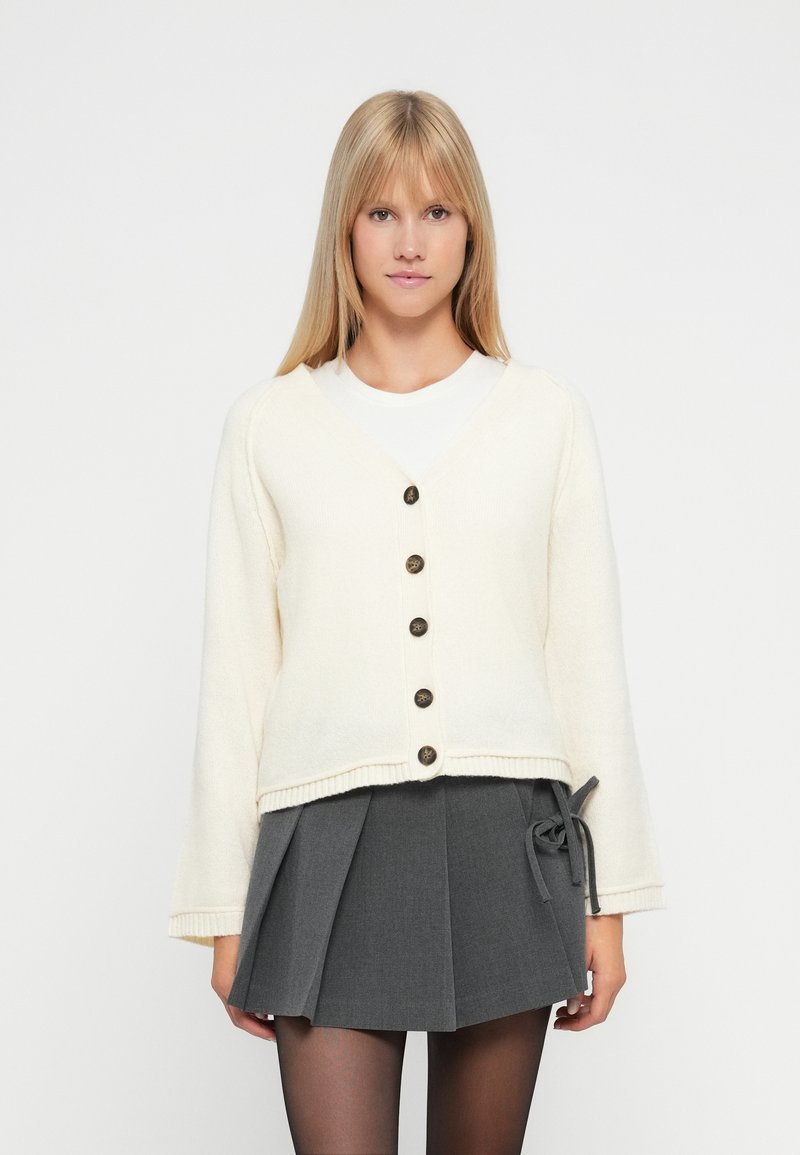 Cremfarbener Cardigan mit V-Ausschnitt, fünf braunen Knöpfen und Glockenärmeln, kombiniert mit einem hoch taillierten grauen Plisseerock. Glatte Textur.