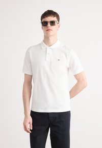 Hvit polo skjorte i myk stoff, med klassisk krave, to-knapps placket og en liten logo på brystet. Brukes med mørke denimjeans.