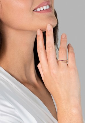 Guldring med pinke ædelstene, glat metaloverflade, rund form, båret på en hånd med naturlige negle, mod en neutral baggrund.