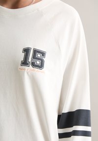 Cremefarbener Sweatshirt mit Raglanärmeln, ausgestattet mit einem marineblauen "15"-Logo und besticktem Text. Marineblaue und weiße Streifen an den Ärmeln. Weicher Stoff.