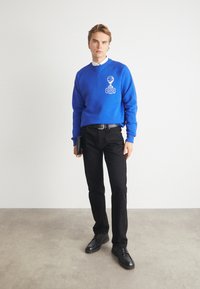 Blauwe sweatshirt met ronde kraag en wit logo, gecombineerd met zwarte broek en zwarte schoenen, bijeengehouden door een zwarte riem en een clutch.