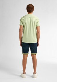 Camiseta verde clara de manga corta, combinada con shorts azul oscuro con dobladillos enrollados y acentos amarillos. Calzado informal completa el conjunto.