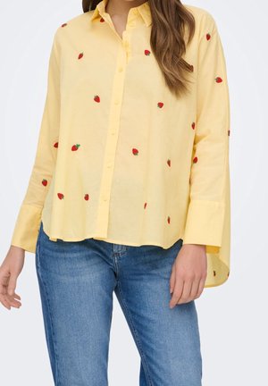Chemise jaune à boutons avec des motifs de fraises rouges portée avec un jean bleu sur une personne aux cheveux bruns.