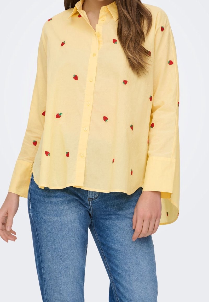 Chemise jaune à boutons avec des motifs de fraises rouges portée avec un jean bleu sur une personne aux cheveux bruns.