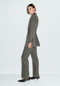 Donna in piedi di lato, vestita con un completo grigio su misura con un blazer sagomato e pantaloni dritti, abbinato a scarpe nere con tacco basso.
