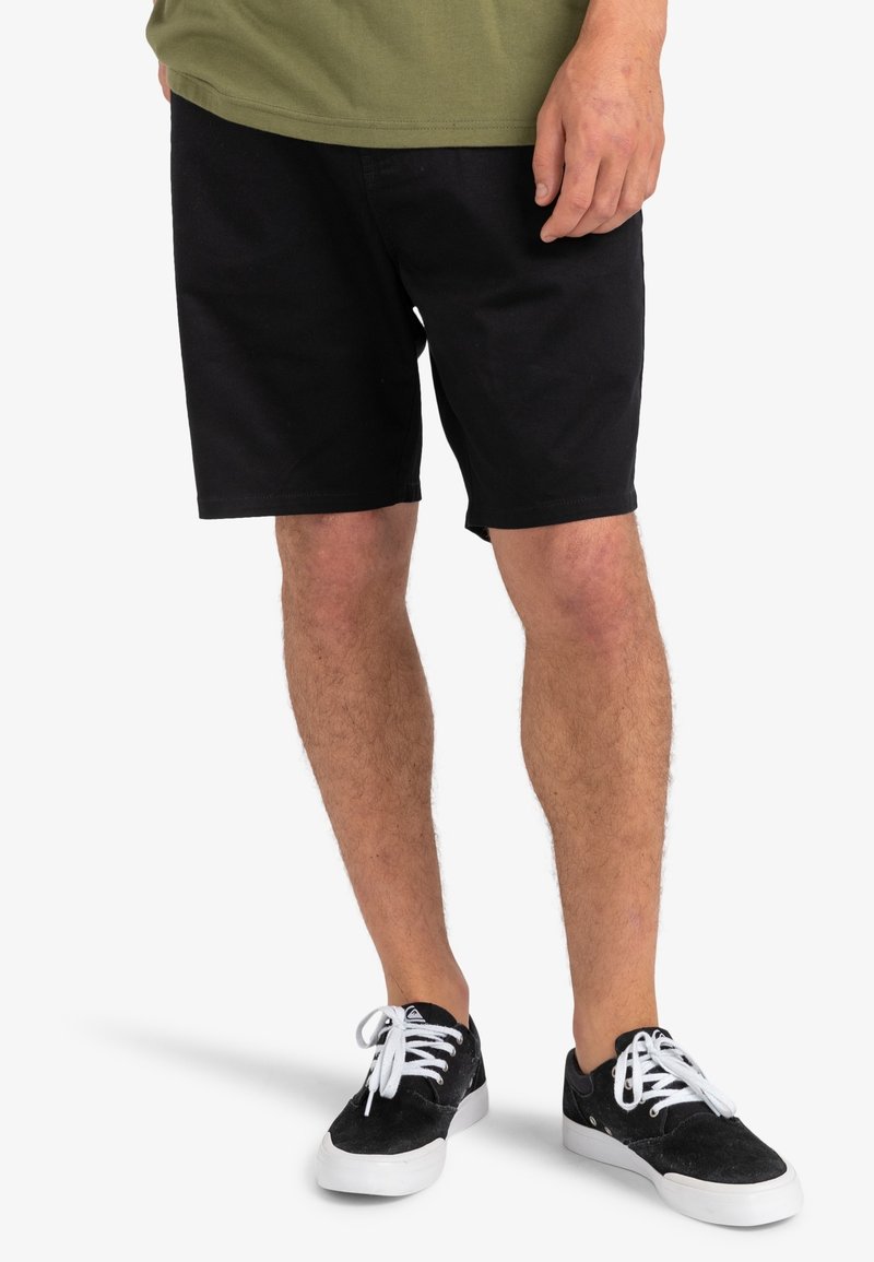 Shorts en coton noir à coupe droite et texture lisse, assortis à des baskets en daim noir avec des lacets et semelles blanches.
