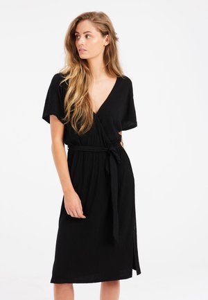 Robe noire en tissu texturé. Elle présente des manches courtes, un décolleté en V, une ceinture nouée à la taille et une longueur mi-cuisse avec une fente sur le côté.
