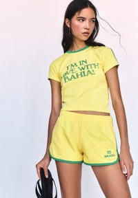 Geel shirt met korte mouwen en groene randen, met de tekst "IK BEN VERLIEFD OP BAHIA!"; bijpassende gele shorts met groene accenten en het logo "BRAZILIË".