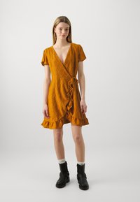 Robe enveloppante jaune avec un petit ourlet volanté, motif en diamants texturé, manches courtes et détail à nouer à la taille. Portée avec des bottines noires.