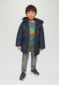 Marineblauwe gewatteerde jas met een capuchon, gecombineerd met een groene graphic t-shirt en grijze jeans. Gekenmerkt door sneakers met zwarte en groene accenten.