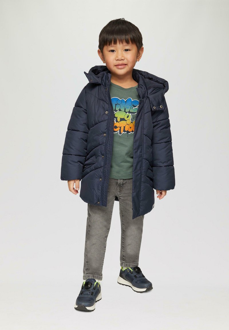 Marineblauwe gewatteerde jas met een capuchon, gecombineerd met een groene graphic t-shirt en grijze jeans. Gekenmerkt door sneakers met zwarte en groene accenten.