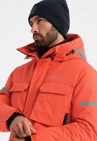 Felgekleurde oranje parka met een ritssluiting aan de voorkant, meerdere zakken en grijze accenten. Bevat een capuchon en ribgebreide manchetten, gemaakt van een gestructureerde stof.
