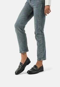 Mocassini in pelle nera con suola piatta, dotati di un accento in metallo argentato, indossati con jeans di denim azzurro chiaro. Dettagli della superficie texturizzati visibili.