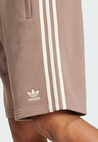 Shorts en coton marron avec trois bandes blanches sur le côté et un petit logo Adidas blanc sur l'ourlet. Dotés de poches et d'une coupe décontractée.