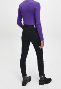 Jeans negros ajustados con bolsillos traseros, de cintura alta, combinados con un suéter de canalé morado. El conjunto complementa unas botas negras hasta el tobillo con cremalleras.