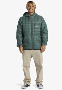Green quilted puffer jacket with a front zip, hood, and a small chest pocket, worn with beige pants and black sneakers.