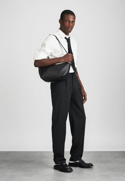 Fekete bőr válltáska sima tapintással, ívelt formával és állítható pánttal. A modellen fehér ing, fekete nyakkendő és fazonra szabott nadrág van.